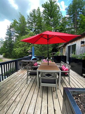 A wooden deck with a table and chairs, a red umbrella, and a view of the surrounding trees. - Willow Cabin - North Frontenac Lodge (Ompah)