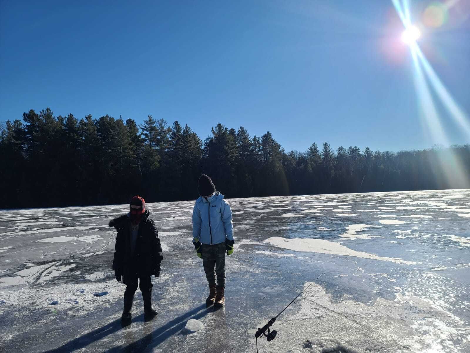 Deux personnes se tiennent sur une surface gelée, une canne à pêche à la main, entourées d’arbres et sous un ciel dégagé.