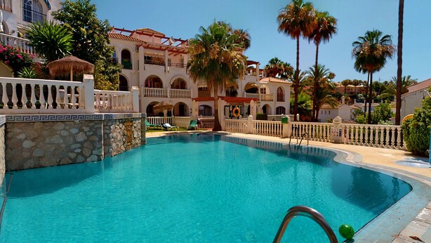 A pool area with a stone wall, palm trees, and a building with arched windows and balconies. - Balcón del Golf - Riviera Cozy Apartment (Mijas Costa)