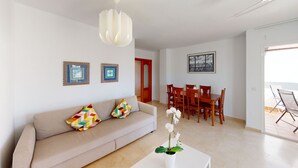 A living room with a beige sofa, colorful pillows, a white table with an orchid, a dining area with wooden chairs, and a framed cityscape picture on the wall. - Balcón del Golf - Riviera Cozy Apartment (Mijas Costa)