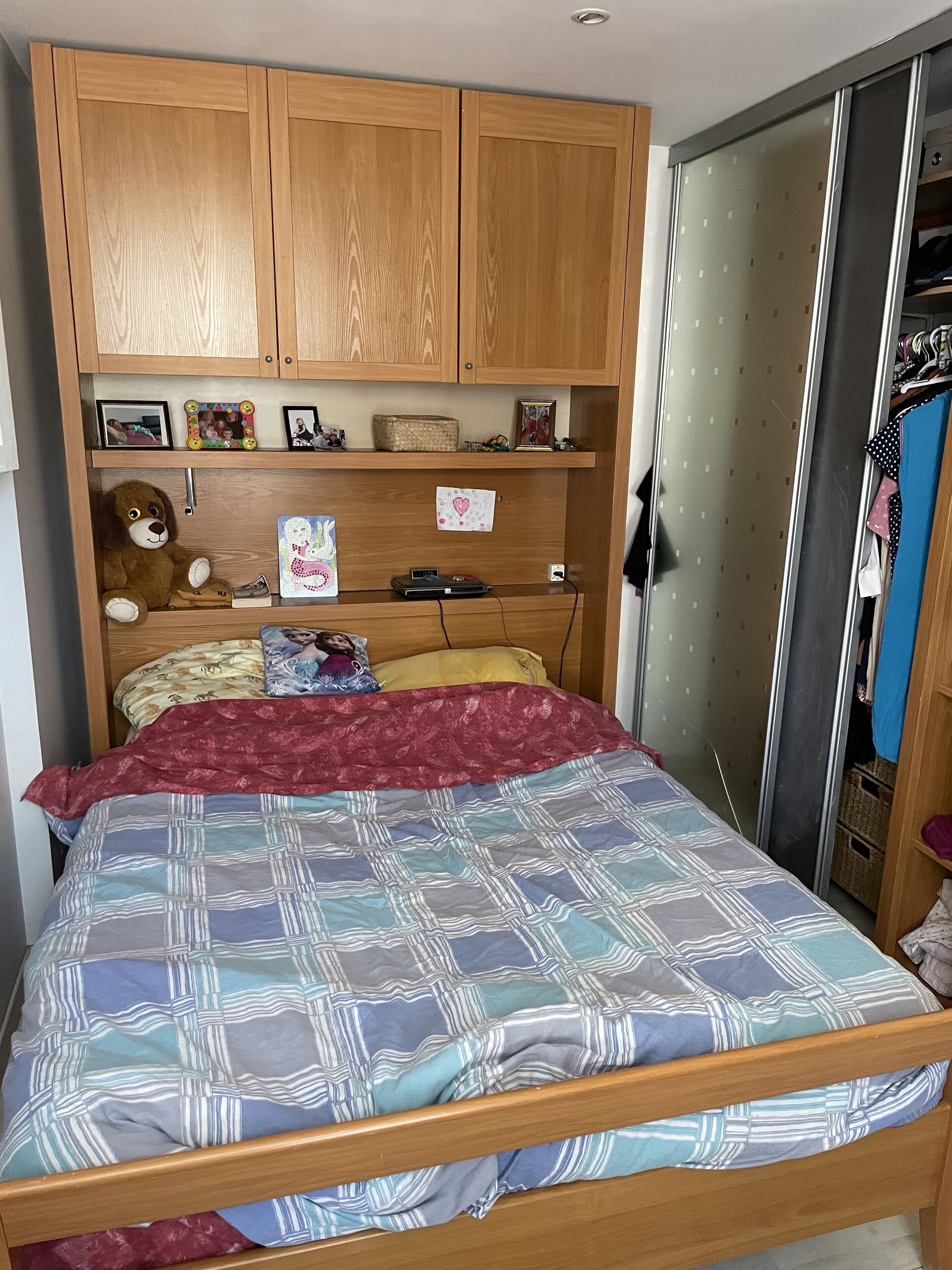 A bedroom with a wooden bed frame, a red and blue patterned bedspread, a teddy bear, and wooden cabinets above the bed.