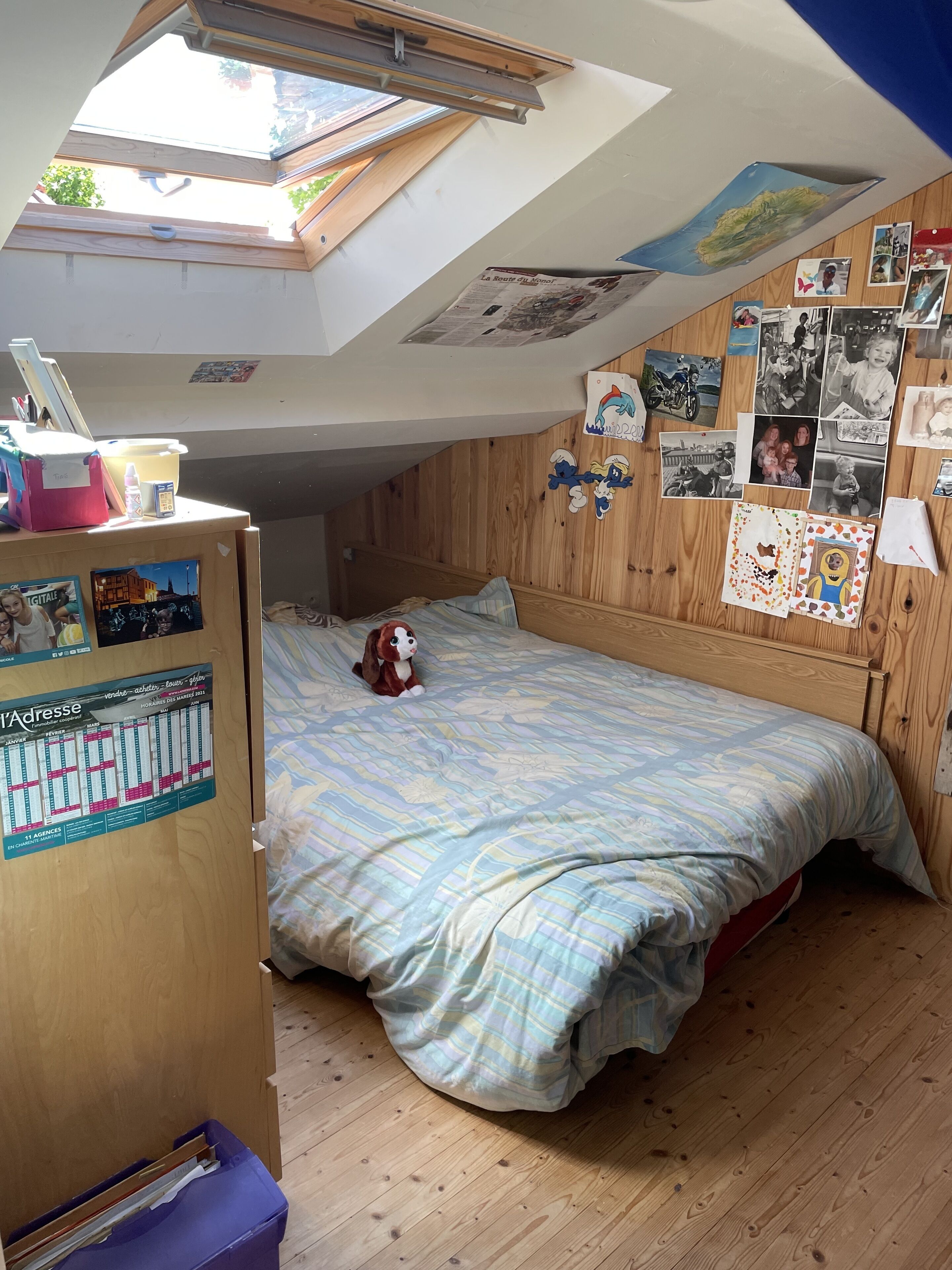 Une chambre avec un plafond en pente, des murs en bois, un lit recouvert d’une couverture à rayures, une petite table avec une lampe et un mur décoré de photos et d’affiches.