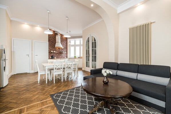 A spacious living area with a dark grey sofa, a round coffee table, and a patterned rug. There is a dining area with white furniture and a brick accent wall. - Native Apartments Batorego 7 (Kraków)