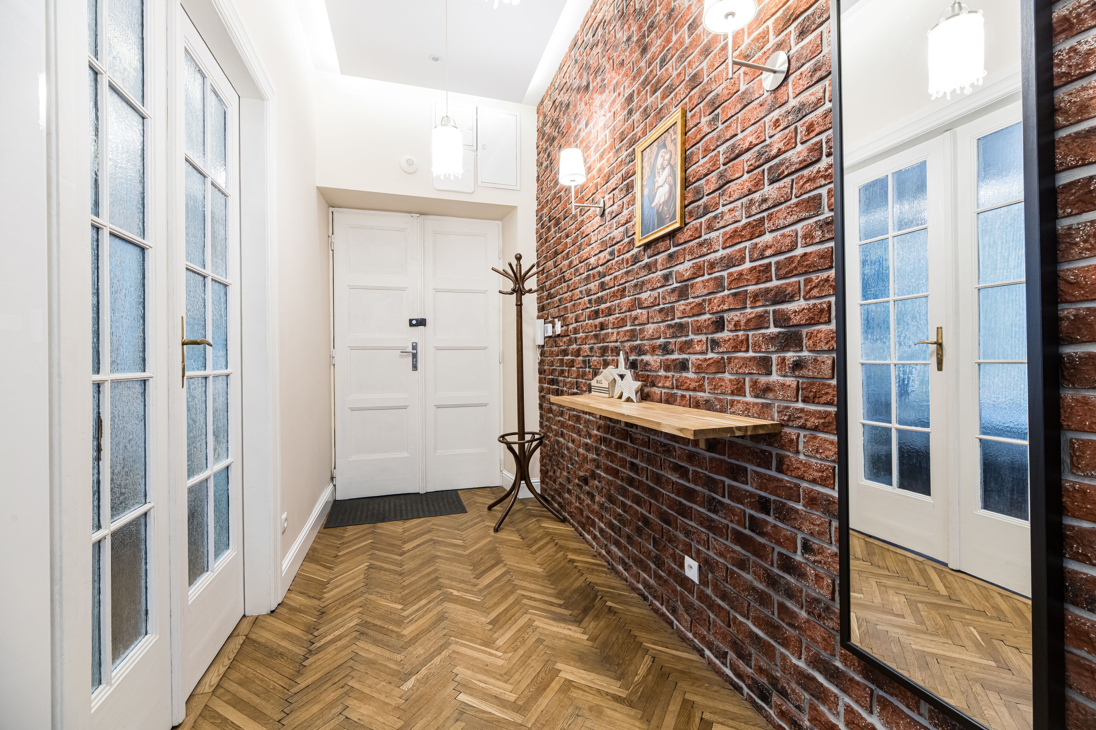 A hallway with a herringbone wooden floor, white doors, a brick wall, and a shelf with decorative items.