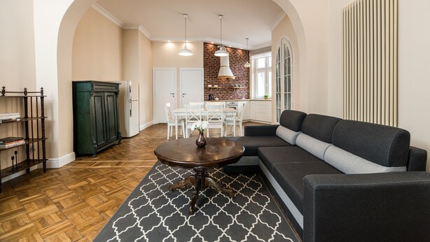 A modern living room with a sectional sofa, a round coffee table, and a patterned rug. There is a dining area with a white table and chairs, and a kitchen with a brick wall and white cabinets. - Native Apartments Batorego 7 (Kraków)
