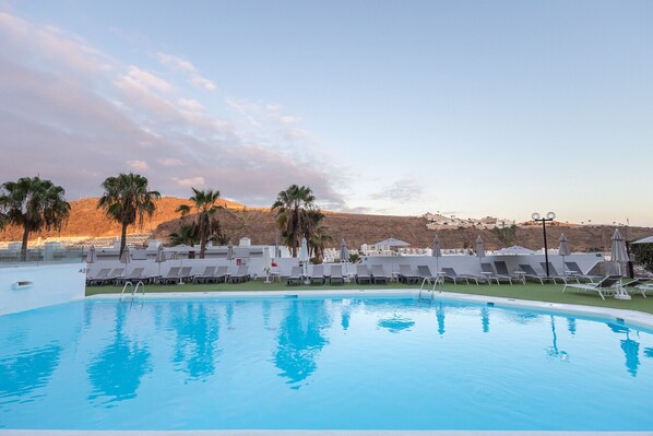A swimming pool with lounge chairs and umbrellas, surrounded by palm trees and a view of a hilly landscape.