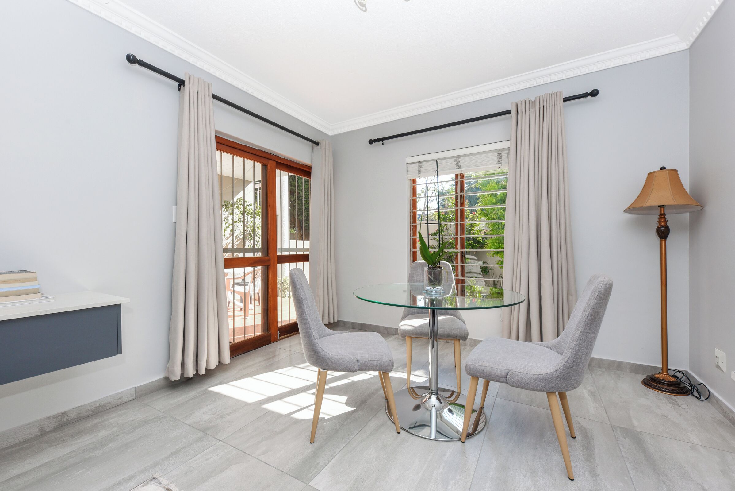 Uma área de jantar moderna com mesa de vidro, duas cadeiras cinzas, um abajur e vista para o verde pela janela.