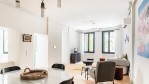 Amplia sala de estar con mesa de comedor, sillas, sofá y televisor. Grandes ventanales ofrecen luz natural y vistas al exterior, con vegetación.