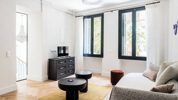 Una sala de estar moderna con un sofá, un televisor sobre un mueble negro, dos mesitas de noche negras y un ventanal con cortinas.