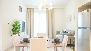 A modern dining area with a round table, white chairs, and a chandelier. There is a sofa and a refrigerator in the background. - Yogi - Elegant Apartment With Balcony Amidst Vibrant Area (Dubai)