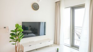A modern living room with a flat-screen TV, a white entertainment unit, a large window with curtains, and a plant. - Yogi - Elegant Apartment With Balcony Amidst Vibrant Area (Dubai)