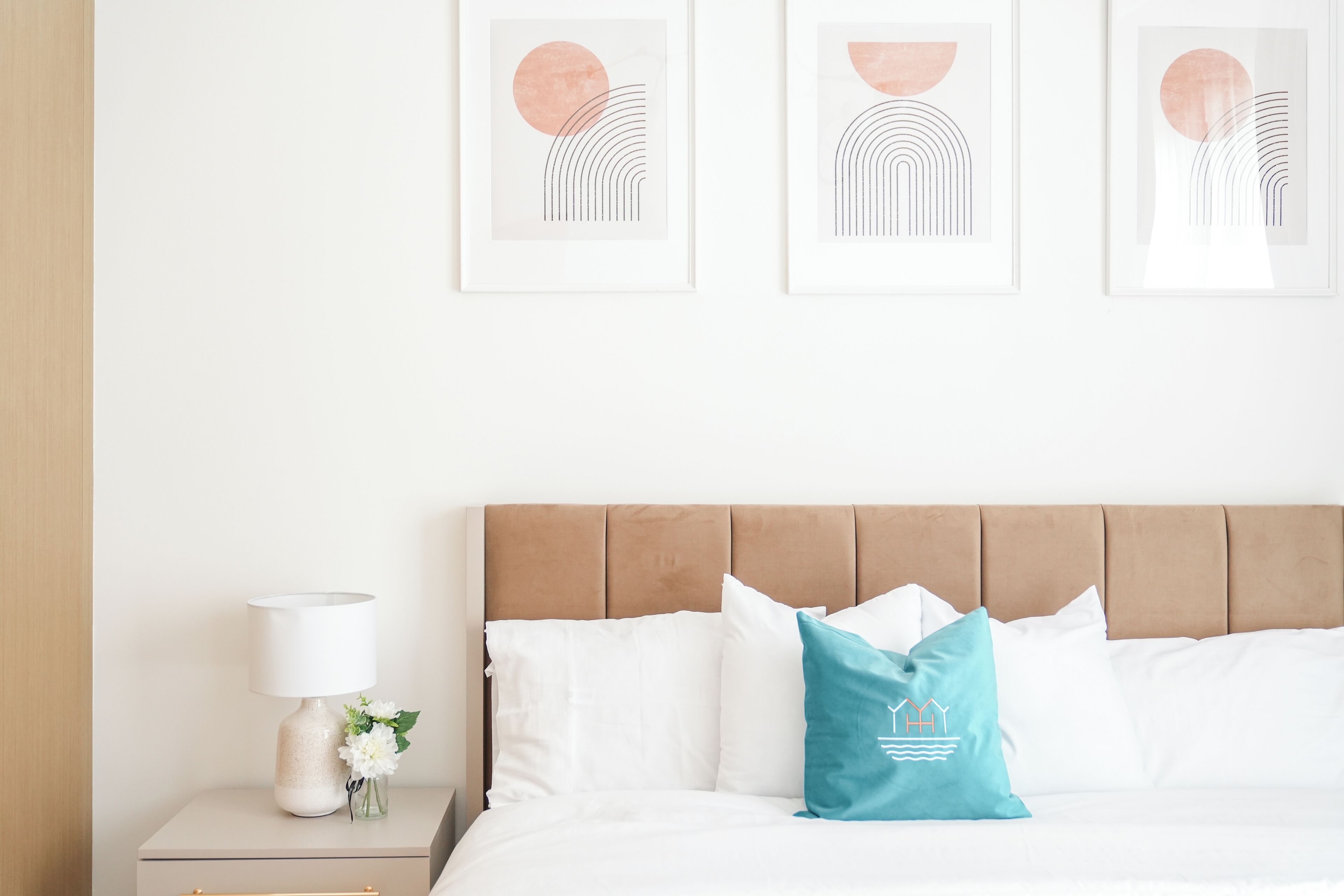 Een keurig opgemaakt bed met witte en teal kussens, een beige hoofdbord, en een nachtkastje met een lamp en een bloemenvaas.