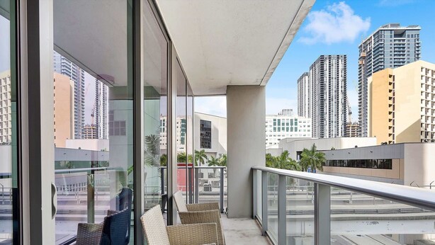 A balcony with wicker furniture, a glass railing, and a view of high-rise buildings.