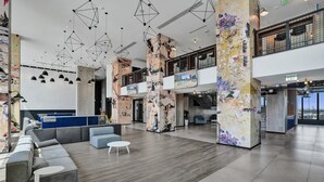 Un hall d’entrée spacieux au design moderne, comprenant un espace salon avec des canapés et une table basse, un plafond haut orné de luminaires aux formes géométriques et un grand mur décoré d’une fresque représentant un paysage urbain.