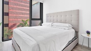A modern bedroom with a large bed, a view of a brick building, and a small table with a plant.