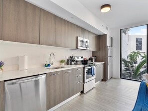 A modern kitchen with stainless steel appliances, wooden cabinets, and a large window offering a view of palm trees. - X Lounge III-Modern Corner Residence (Miami)