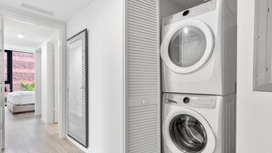 A laundry room with two stacked washing machines, a large mirror, and a bed in the adjacent room.