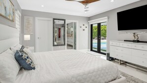 Quarto moderno com uma cama grande, ventilador de teto, TV e vista para uma piscina através de portas de vidro.