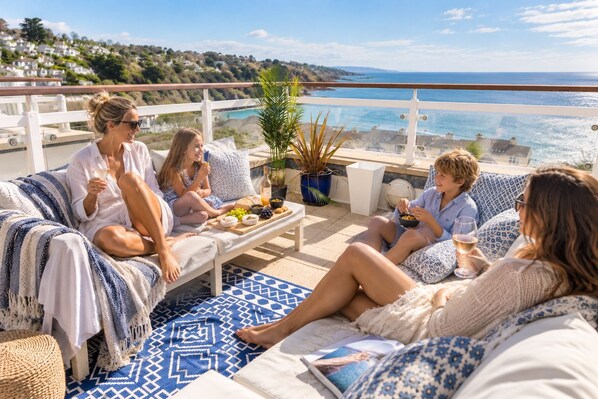 A group of people sitting on a balcony with a view of the sea, a table with food and drinks, and a potted plant.