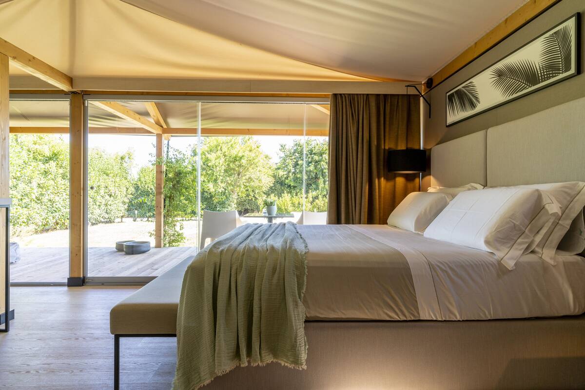 A bedroom with a large bed, a view of greenery outside, a table with chairs, and a ceiling fan.