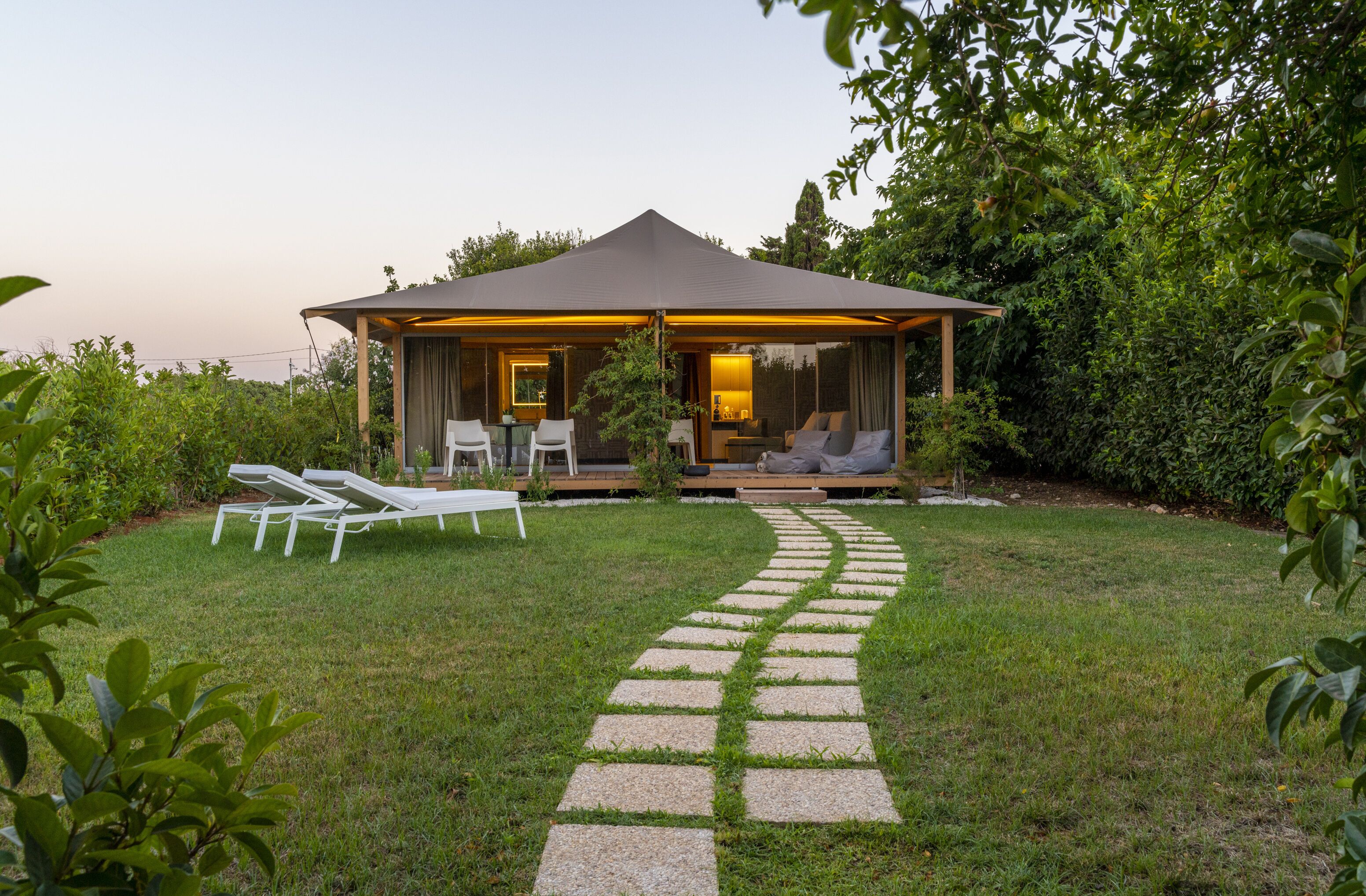 A cozy outdoor lounge area with a covered seating space, a stone pathway leading to it, and lush greenery surrounding the area.