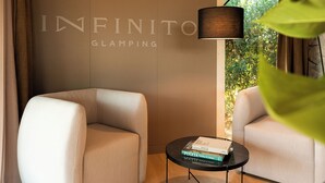 A modern room with a large wall-mounted sign reading "INFINITO GLAMPING," two plush armchairs, a small round table with a lamp, and a window with greenery outside.