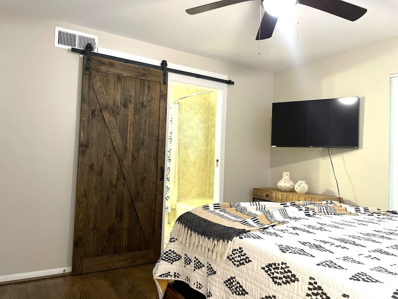 A bedroom with a wooden sliding door, a bed with patterned bedding, a flat-screen TV, and a ceiling fan.