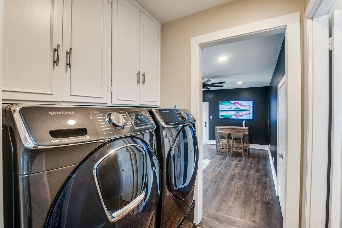 A laundry room with two Samsung SuperSpeed Steam Vortex washers, a wooden desk, and a TV in the adjacent room.
