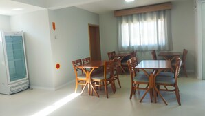 A dining area with wooden tables and chairs, a refrigerator, and a window with curtains. - Pousada Ametista (Penha)