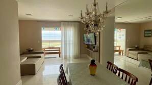 A spacious dining area with a long table, chairs, and a chandelier. There is a sofa and a television in the background. - 1101 Ap Vista mar atrás da passarela (Aracaju)