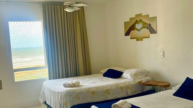 A hotel room with two beds, a large window with curtains, a ceiling fan, and a wall art piece. - 1101 Ap Vista mar atrás da passarela (Aracaju)