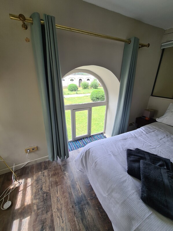 Ein Schlafzimmer mit einem großen Bett, einem Fenster mit Vorhängen, Blick auf einen Garten und Holzboden.