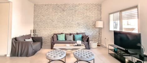 A living room with a brick wall, a sofa, two ottomans, a coffee table, a TV, and a lamp.