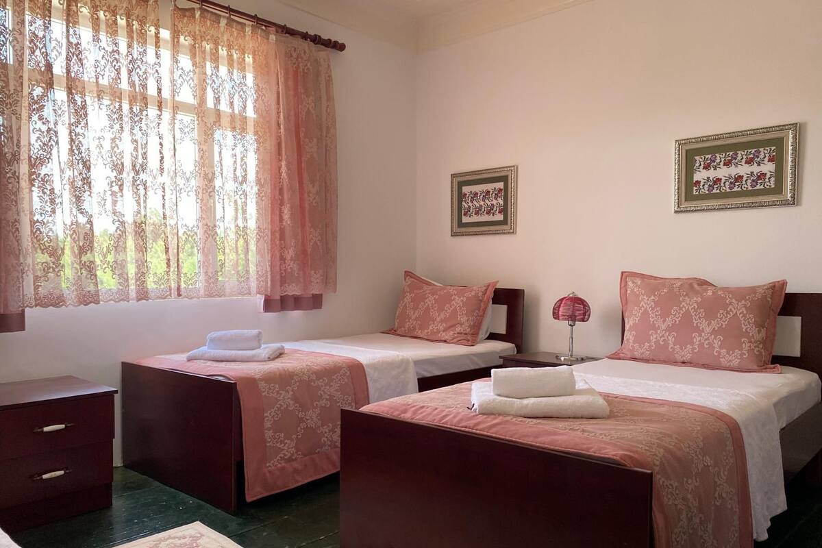A hotel room with two single beds, a nightstand with a lamp, a dresser, and a window with sheer curtains.