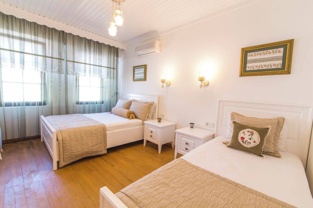 A hotel room with two beds, a wooden floor, a window with curtains, an air conditioner, and wall-mounted lights.