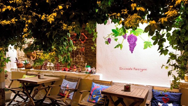 An outdoor seating area with wooden tables and chairs, surrounded by potted plants and a stone wall. - Bozcaada Butik Pansiyon (Bozcaada)