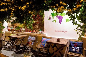 Un espacio al aire libre con mesas y sillas de madera, rodeado de macetas con plantas y un muro de piedra.