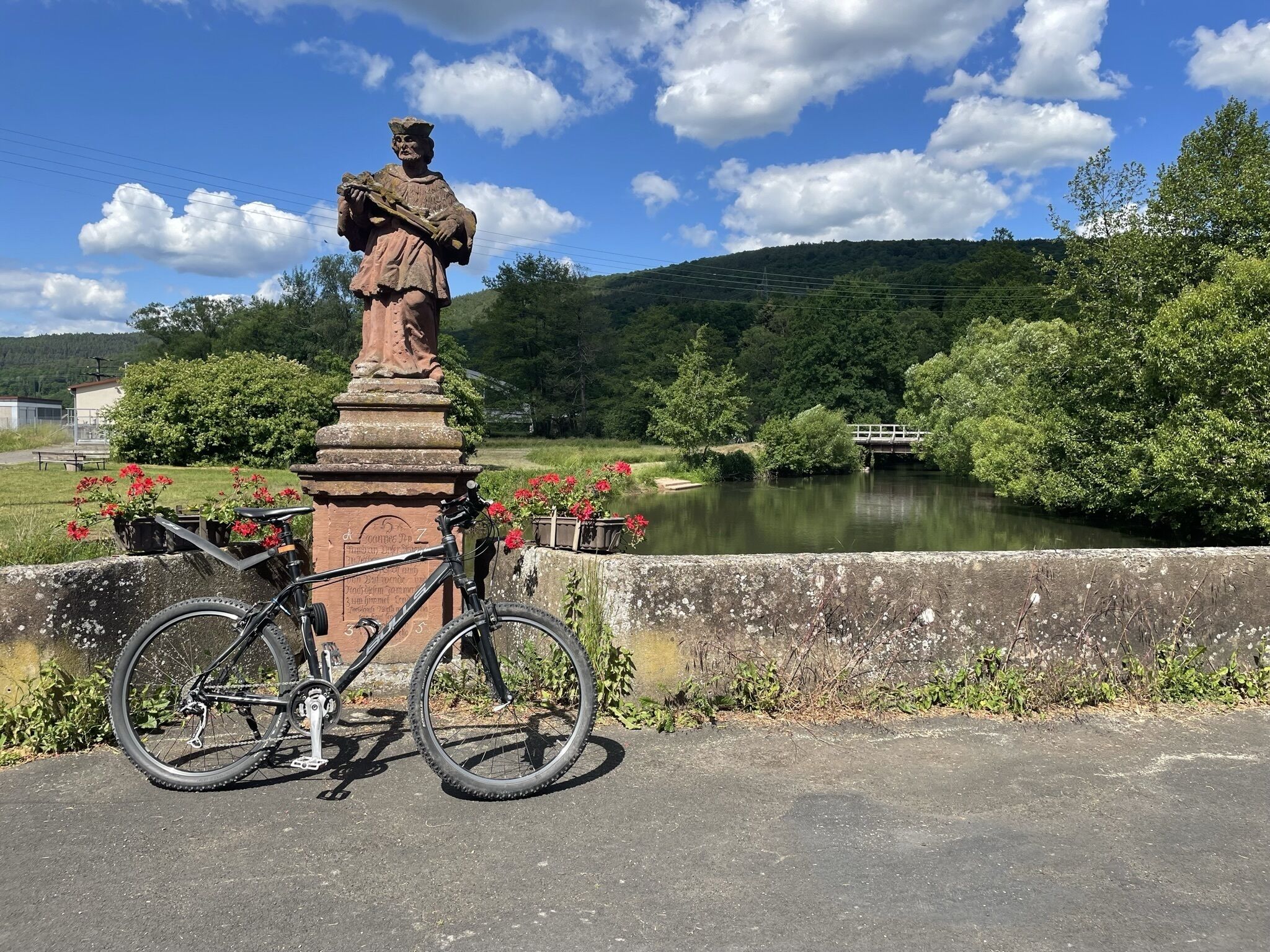 A statue on a pedestal with a bicycle parked nearby, set against a backdrop of a river and greenery.