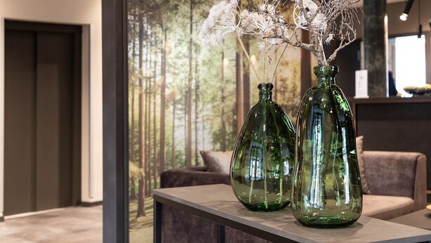 A modern interior with two green glass vases on a table, a large window with a forest view, and a dark door. - Hotel Imhof (Gemuenden)