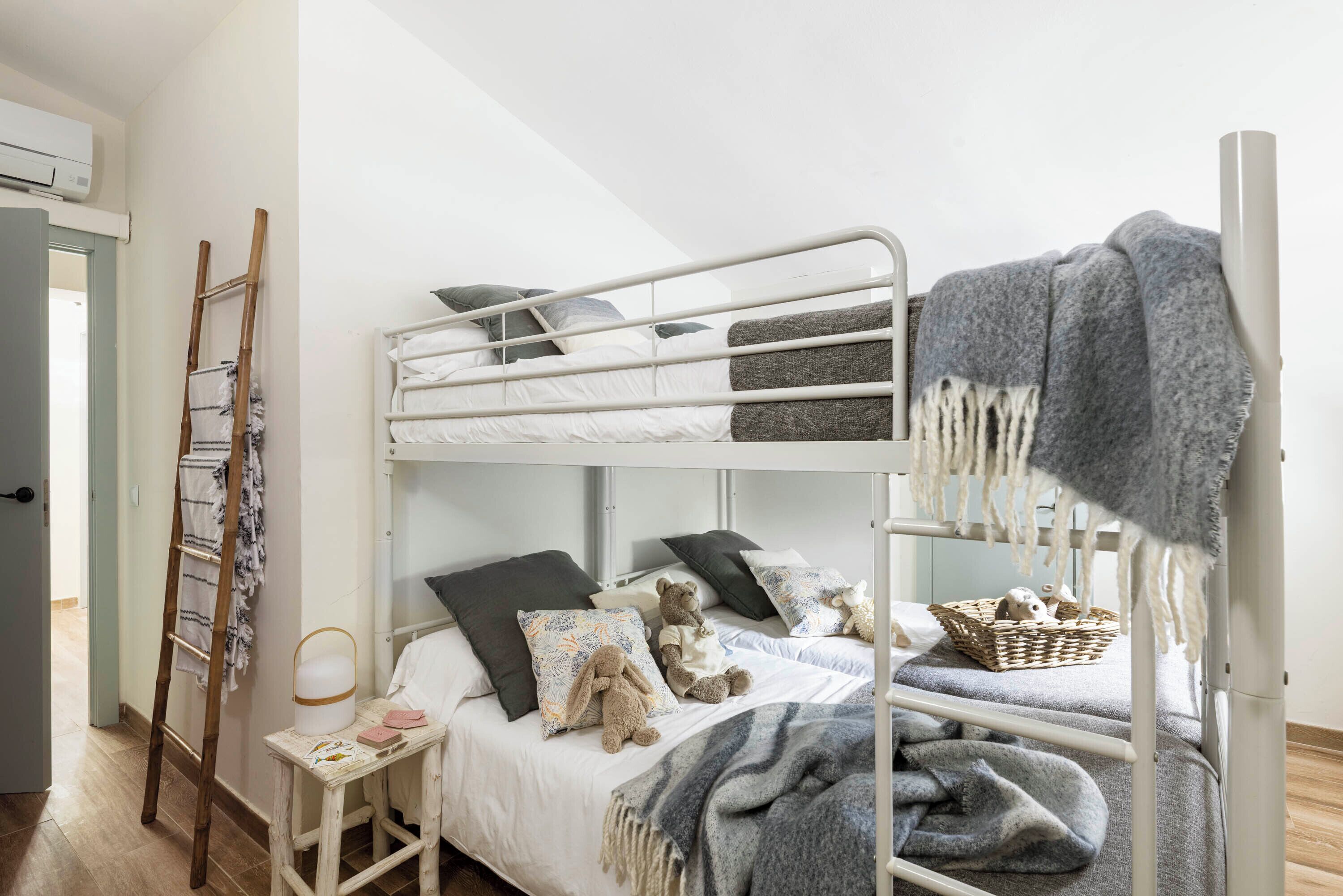 Una cama litera con escalera, una mesita de noche con lámpara, y una cesta en la litera inferior.