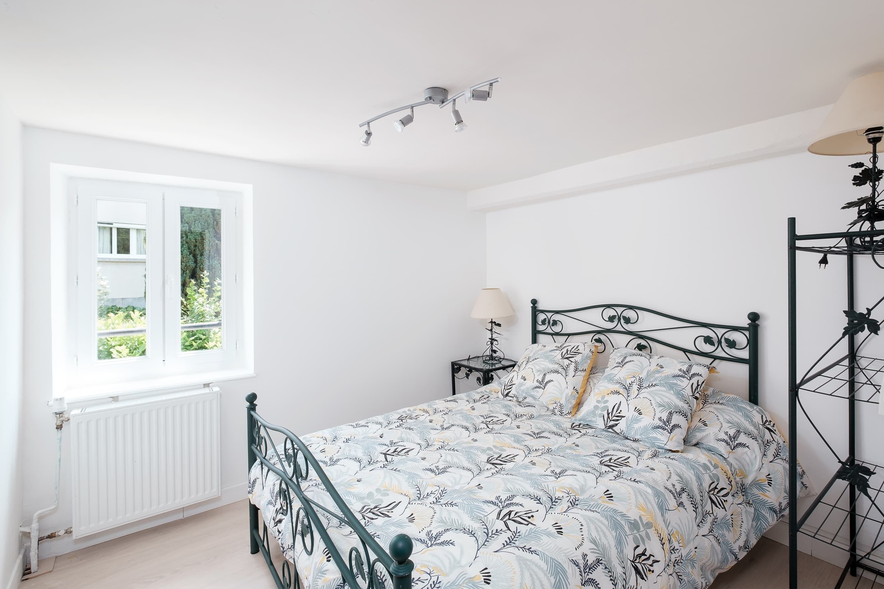 A bedroom with a bed, a window, a radiator, a bedside table with a lamp, and a floor lamp.