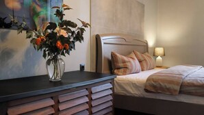 Un dormitorio con cama, mesita de noche, lámpara y un florero con flores sobre una cómoda.