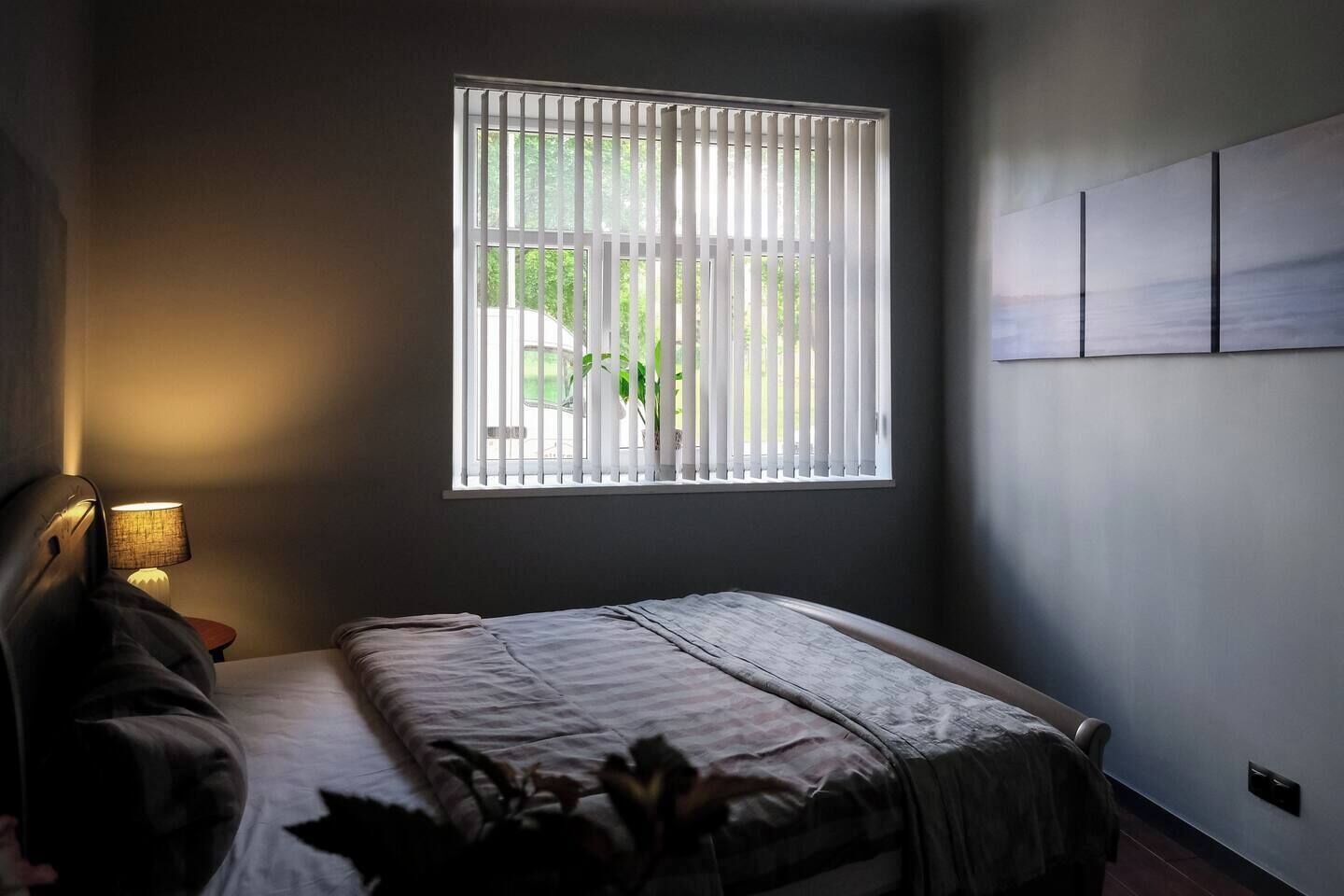 Una camera da letto con un letto, un comodino, una lampada, una finestra con tende e un punto luce a parete.