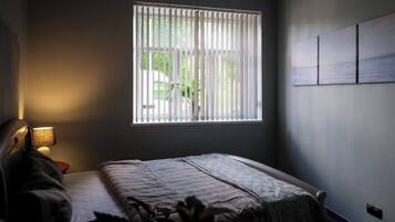Una camera da letto con un letto, un comodino, una lampada, una finestra con tende e un punto luce a parete.