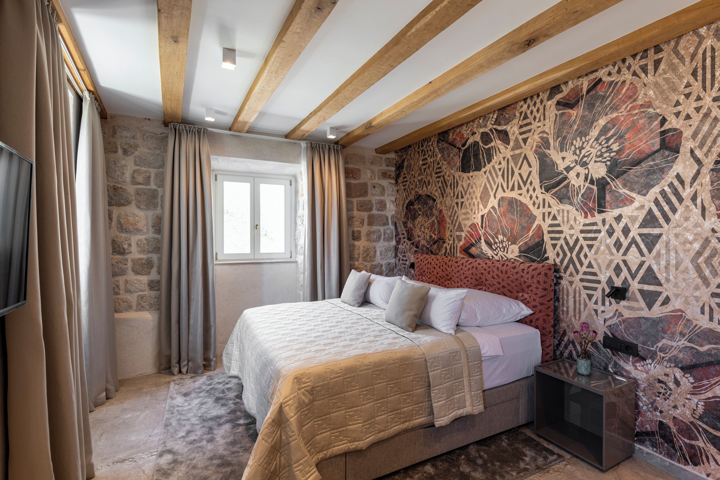 A bedroom with a stone wall, a bed with a patterned headboard, and a flat-screen TV on the left.