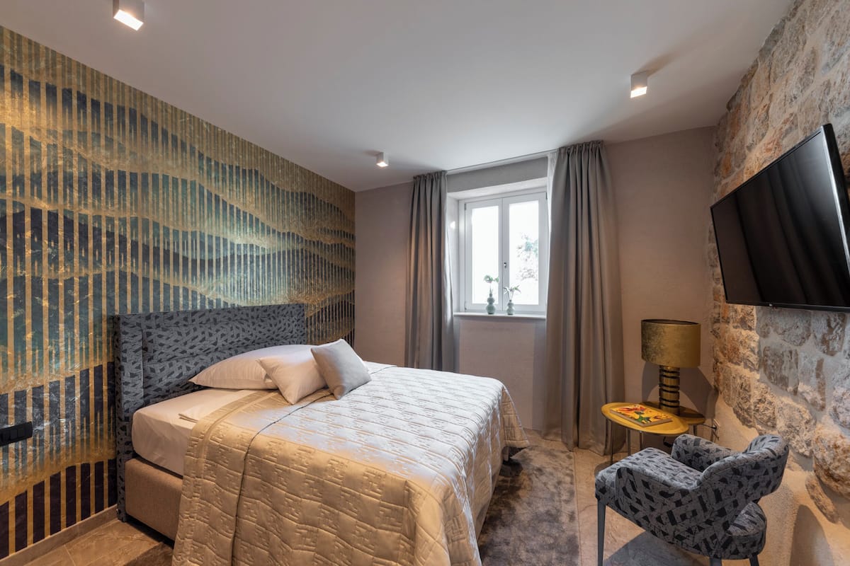 A bedroom with a bed, a chair, a TV, a window with curtains, and a lamp.
