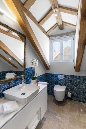 A modern bathroom with a large mirror, white vanity, and a white toilet. The ceiling features exposed wooden beams, and the walls have a blue geometric tile pattern.