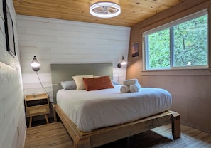 A bedroom with a wooden bed, white bedding, two bedside lamps, a nightstand, a window with a view of trees, and a ceiling fan. - Unbelievably Scenic Mountainside Cabin (Irwin)