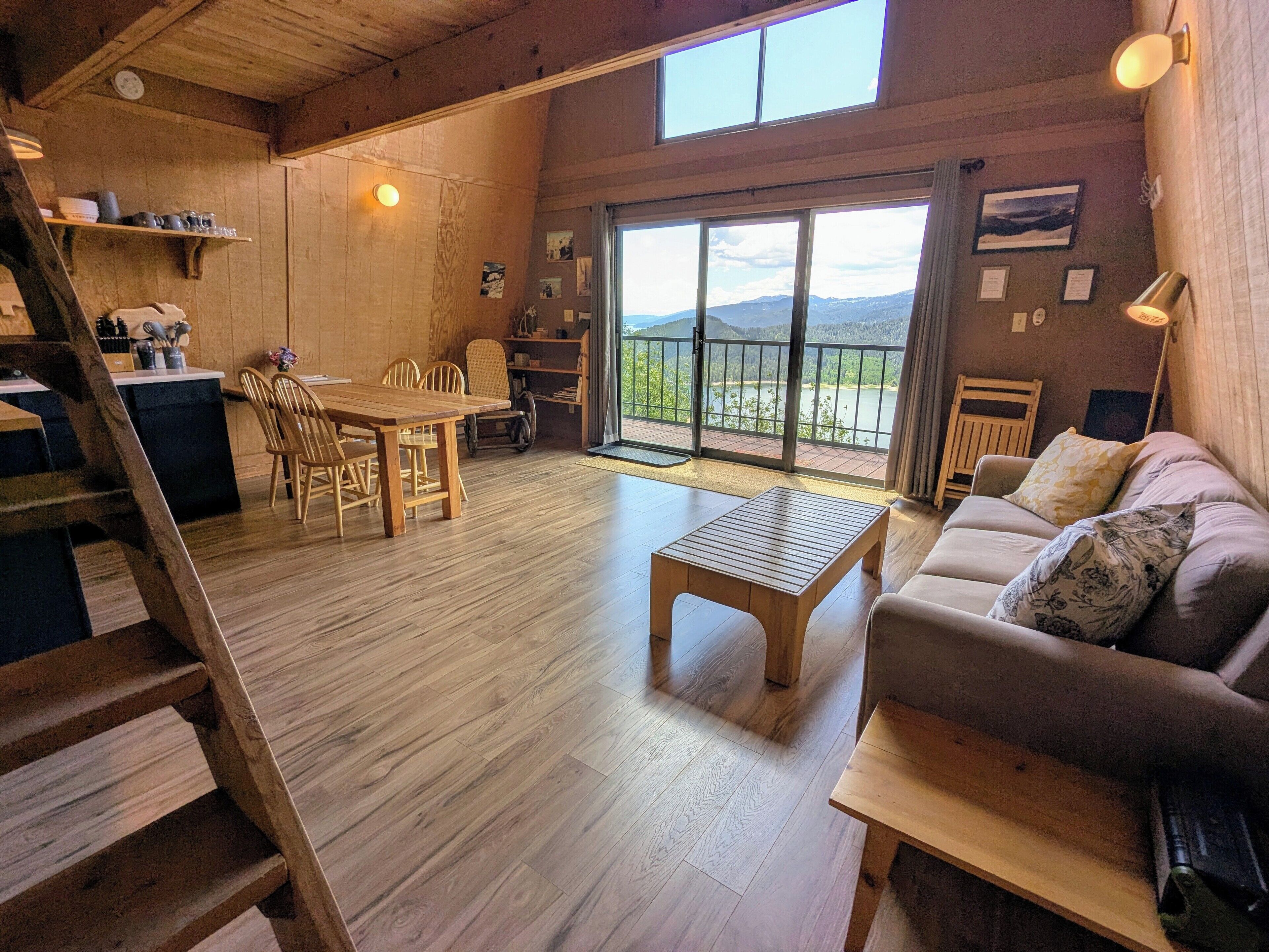 A spacious living area with a wooden floor, a dining table, a sofa, and a balcony with a view of the mountains.