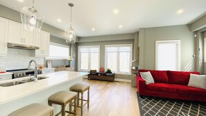 A spacious living area with a red sofa, wooden bar stools, and a kitchen with white cabinets and a stainless steel range hood. - Near Ridley College  (St. Catharines)